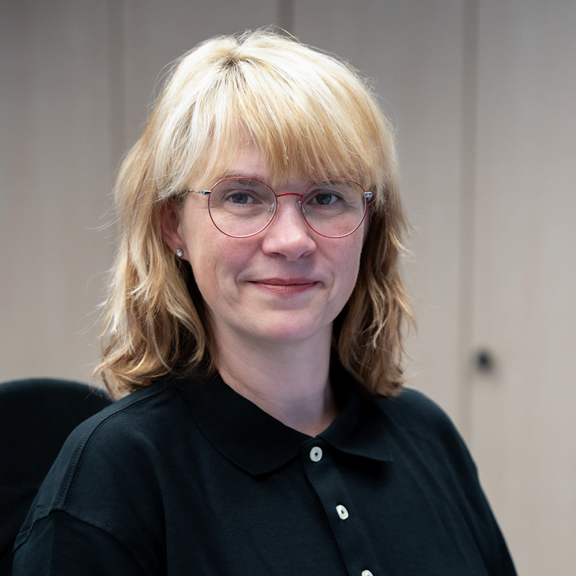 Eine Frau mit blonden Haaren und Brille lächelt sanft und trägt ein schwarzes Hemd