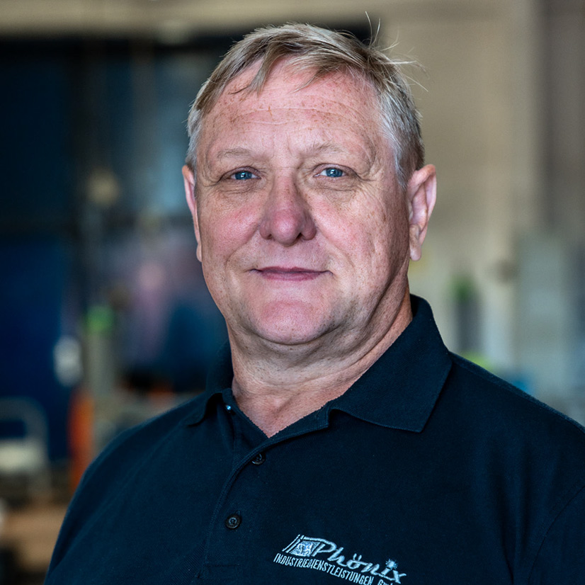 Eine ältere Person mit kurzem blondem Haar und einem nachdenklichen Blick trägt ein dunkles Polohemd mit einem Firmenlogo.