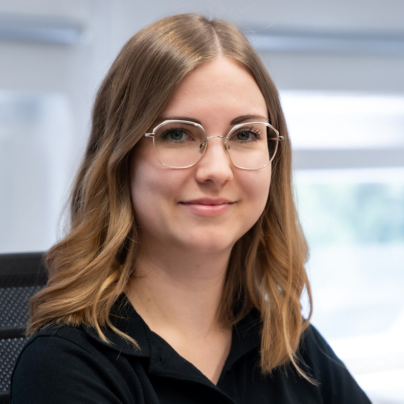 Junge Frau mit Brille lächelt sanft, braunes Haar und schwarzes Oberteil.