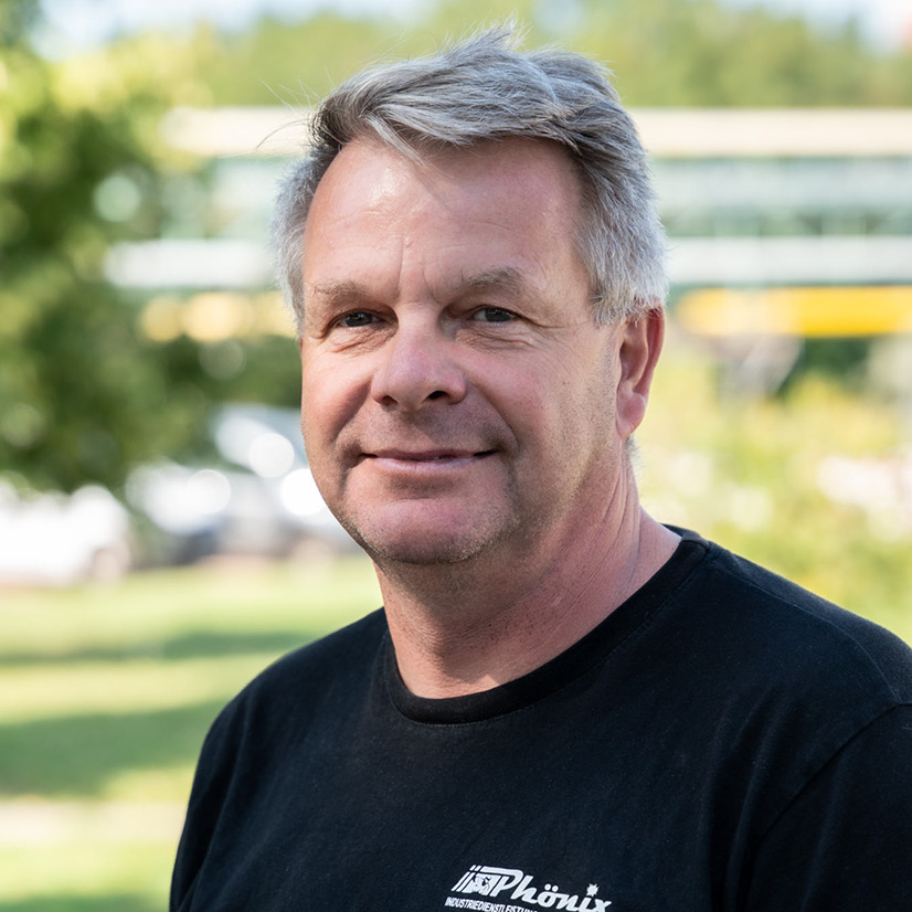 Ein lächelnder älterer Mann mit grauem Haar trägt ein schwarzes T-Shirt im Freien