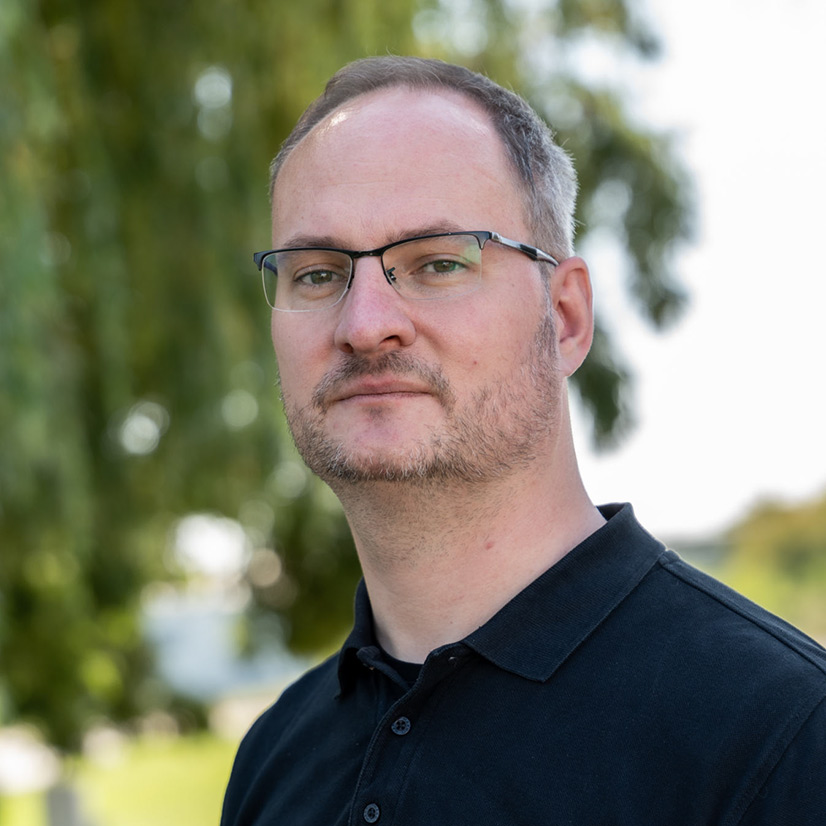 Ein Mann mit Brille und Bart schaut ernst, trägt ein schwarzes Hemd draußen